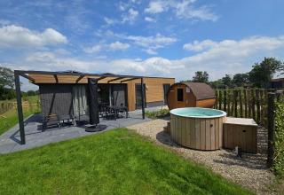 Vista exterior de una cabaña moderna con terraza, muebles de jardín, sauna de barril y jacuzzi redondo.