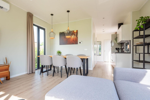 Moderne lodge met eettafel, zes grijze stoelen, open keuken en zonlicht dat naar binnen valt.