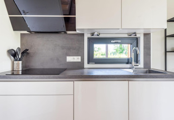 Moderne keuken met witte kasten, spoelbak, kookplaat en raam met uitzicht op de natuur.