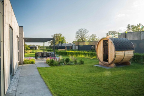 Lodge moderno con sauna de madera en el jardín, comedor exterior y vegetación bajo un día soleado.