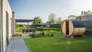 Lodge moderno con sauna de madera en el jardín, comedor exterior y vegetación bajo un día soleado.