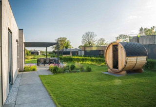 Lodge moderno con sauna de madera en el jardín, comedor exterior y vegetación bajo un día soleado.