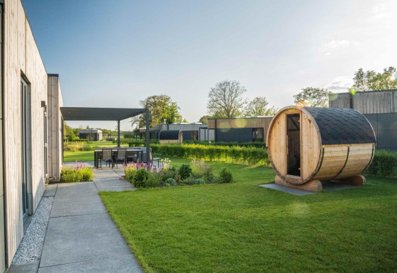Lodge moderne avec sauna en bois dans le jardin, coin repas extérieur et verdure par une journée ensoleillée.