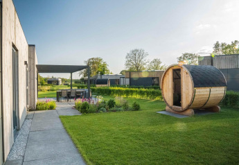 Lodge moderno con sauna de madera en el jardín, comedor exterior y vegetación bajo un día soleado.