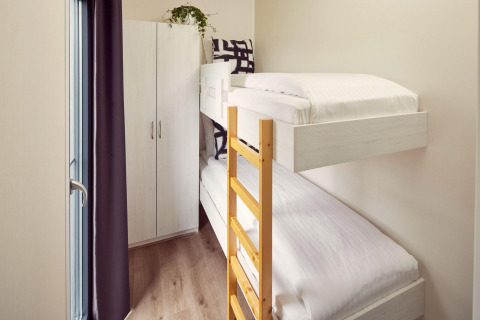Kleine kamer in een lodge met stapelbedden, houten ladder, kast en veel daglicht langs het raam.