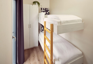 Kleine kamer in een lodge met stapelbedden, houten ladder, kast en veel daglicht langs het raam.