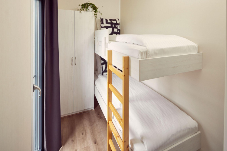 Habitación pequeña en un lodge con literas, escalera de madera, armario y luz natural desde la ventana.