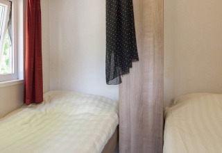 To enkeltsenge i et lille værelse med vindue, rødt gardin og skab på Lodge i Holiday park Witterzomer.