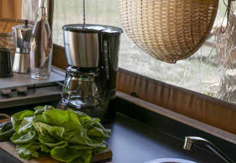 Køkkenområde i en safaritelts campingplads med kaffemaskine, salatblade og fletkurv ved vinduet.