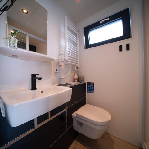 Baño moderno en una casa flotante con inodoro suspendido, lavabo, grifo negro y ventana pequeña.