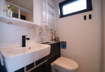 Bagno moderno su una casa galleggiante con wc sospeso, lavabo, specchio, rubinetto nero e finestra.