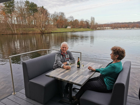 Ældre par sidder ved et bord på Houseboat Marina Mookerplas, med udsigt over sø og træer.