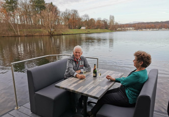 Ældre par sidder ved et bord på Houseboat Marina Mookerplas, med udsigt over sø og træer.