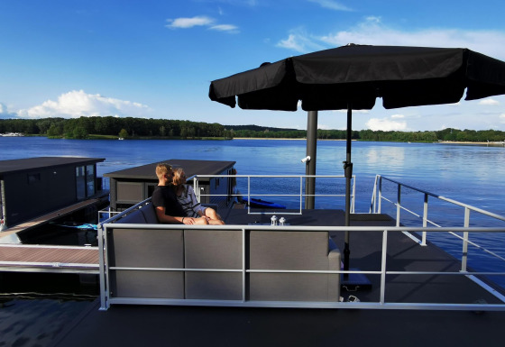 Een stel zit onder een zwart zonnescherm op het terras van een woonboot bij Marina Mookerplas in Nederland.