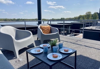 Vista da Houseboat Marina Mookerplas con sedie all’aperto, tazze di caffè e dolci sullo sfondo del lago.