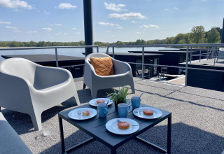 Vista da Houseboat Marina Mookerplas con sedie all’aperto, tazze di caffè e dolci sullo sfondo del lago.