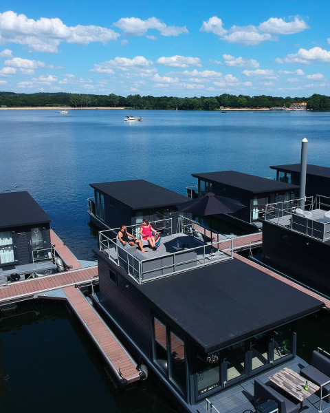 To personer slapper af på taget af en husbåd ved Marina Mookerplas, omgivet af smuk sø og natur.