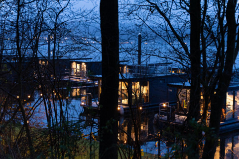 Vista nocturna de casas flotantes iluminadas en Houseboat Marina Mookerplas, rodeadas de árboles en los Países Bajos.
