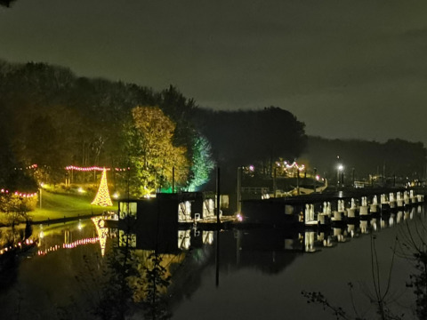 Foto nocturna de una casa flotante en Marina Mookerplas, Países Bajos, con árboles iluminados y reflejos.