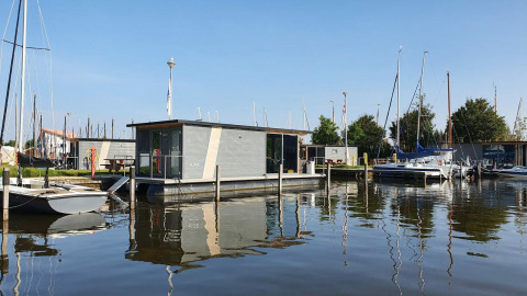 Hausboot am Wasser im Marina Heeg in den Niederlanden, umgeben von Segelbooten und blauem Himmel.