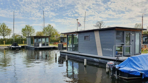 Casa flotante moderna en Marina Heeg, Países Bajos, rodeada de árboles, agua y botes bajo cielo azul.