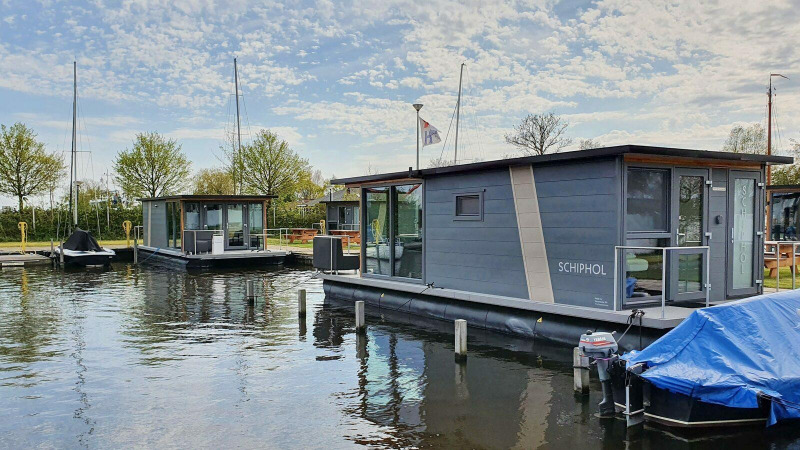 Casa flotante moderna en Marina Heeg, Países Bajos, rodeada de árboles, agua y botes bajo cielo azul.