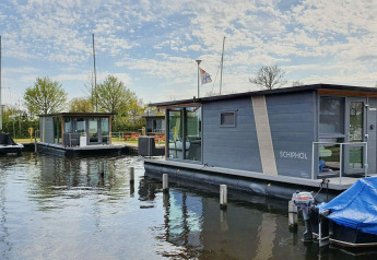 Casa flotante moderna en Marina Heeg, Países Bajos, rodeada de árboles, agua y botes bajo cielo azul.