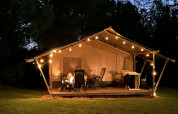Tienda safari con terraza y luces en Camping Emmen, Países Bajos, fotografiada de noche con personas fuera.