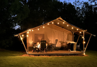 Tienda safari con terraza y luces en Camping Emmen, Países Bajos, fotografiada de noche con personas fuera.