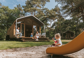 Famiglia rilassata davanti alla Wikkelhouse XL lodge nel bosco, bambini che giocano sullo scivolo.