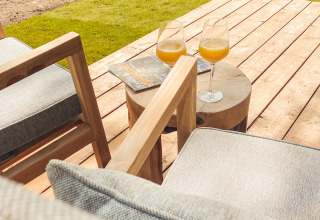 Buitenterras van een lodge met twee glazen sinaasappelsap, houten stoelen, tafeltje en magazines in de zon.