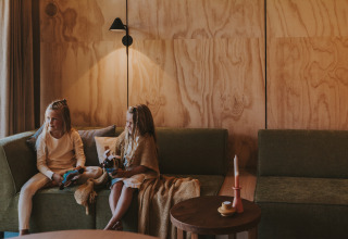 Twee kinderen zitten op groene zetels in een gezellige lodge met houten muren en zachte verlichting.
