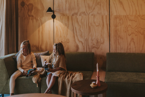 To børn sidder på grønne sofaer i en hyggelig lodge med trævægge og dæmpet belysning.