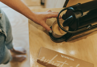Una persona prepara café con una máquina Nespresso negra en una cabaña, con una caja de cápsulas en la mesa.