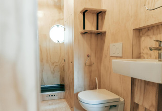Baño moderno con revestimiento de madera, inodoro, lavabo, estantes y ventana redonda en Wikkelhouse XL, Países Bajos.