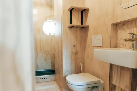 Baño moderno con revestimiento de madera, inodoro, lavabo, estantes y ventana redonda en Wikkelhouse XL, Países Bajos.