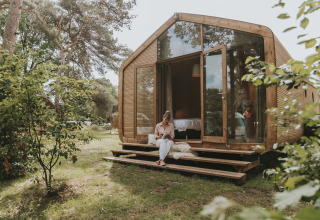 Eine Frau liest ein Buch auf der Holzstufe einer modernen Wikkelhouse XL-Lodge im Grünen in Beerze Bulten, Niederlande.