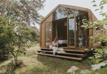 Kvinde læser en bog på trappen foran en moderne Wikkelhouse XL-lodge omgivet af grøn natur i Beerze Bulten, Holland.