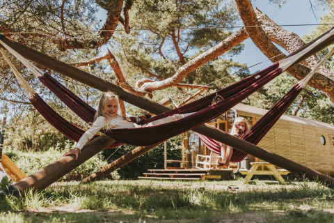 Dos niños juegan en hamacas bajo árboles frente al alojamiento Wikkelhouse XL en Beerze Bulten, Países Bajos.