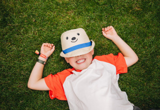 Un bambino sorridente sdraiato sull’erba con un cappello con faccina, fotografato in un glamping.