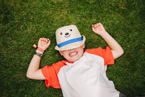 Un bambino sorridente sdraiato sull’erba con un cappello con faccina, fotografato in un glamping.