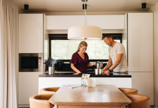 A couple cooking together in the modern kitchen of Hill Lodge at Beerze Bulten, Netherlands, with dining area.