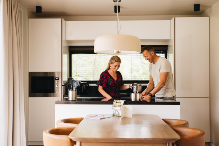 A couple cooking together in the modern kitchen of Hill Lodge at Beerze Bulten, Netherlands, with dining area.