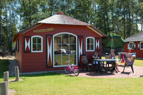 A red nature house with white windows, people sitting outside at a table, surrounded by green lawn.
