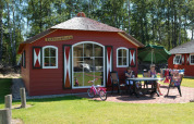 Une maison nature rouge avec fenêtres blanches, des gens assis dehors à une table, entourés de pelouse verte.