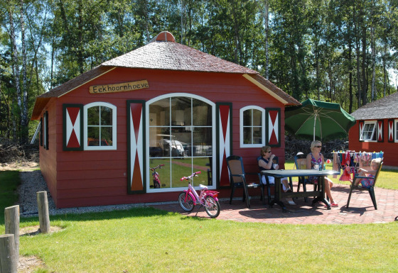 Une maison nature rouge avec fenêtres blanches, des gens assis dehors à une table, entourés de pelouse verte.