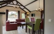 Salon lumineux dans une maison nature avec canapés rouges, chaises vertes et grandes fenêtres sur la nature.