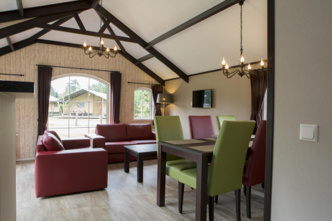 Lichte woonkamer in een natuurhuisje met rode zetels, groene stoelen en grote ramen met zicht op de natuur.