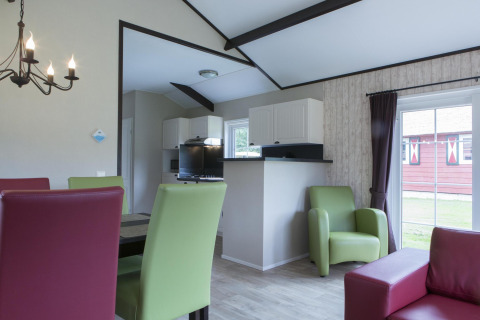 Sala de estar en una Nature house con sillas verdes y rojas, cocina moderna y grandes ventanales.