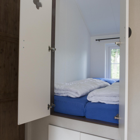 Coin nuit lumineux dans une maison nature avec draps bleus, couettes blanches et fenêtre sur la verdure.